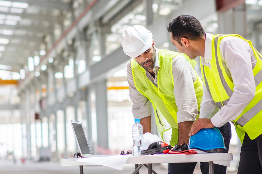 Two construction workers in safety gear discussing plans while reviewing information on a laptop in an industrial setting.